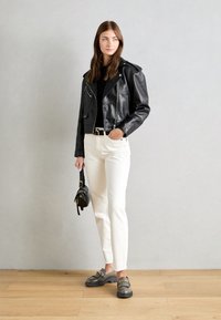 Giacca in pelle nera da motociclista, jeans bianchi a gamba dritta, mocassini grigi con dettagli in catena e una piccola borsa nera. Sfondo semplice.
