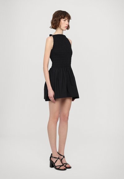 Black sleeveless dress with smocked bodice and flared skirt. Features a high neckline and a bow detail at the back. Worn with heeled sandals.