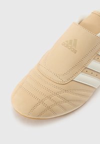 Beige athletic shoe with a smooth leather finish, featuring white striped accents and quilted stitching details, showcasing a minimalist design.