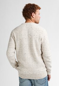 Gebreide trui in lichtbeige met een gestructureerd patroon, ronde halslijn en geribbelde manchetten. Draagt met blauwe jeans.