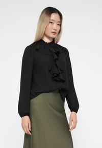 Black blouse with ruffled neckline and front, long sleeves with gathered cuffs, paired with an olive-green skirt, smooth texture.