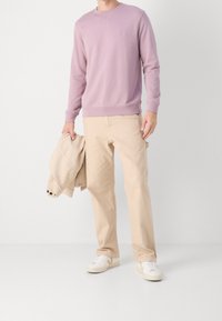 Světle fialová mikina s malým logem, doplněná béžovými kalhotami. Model drží béžovou bundu a má na sobě bílé tenisky s jemnými akcenty.