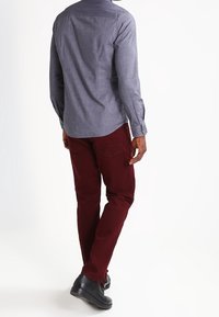 Camicia azzurro chiaro a maniche lunghe con polsini abbottonati, abbinata a pantaloni bordeaux e scarpe nere, mostrata da dietro contro uno sfondo neutro.