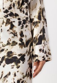 Blusa di seta color crema con stampa astratta di leopardo marrone e nero; presenta una vita con cintura e maniche larghe con polsini. Texture liscia.