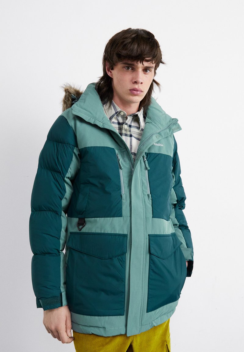 Veste rembourrée en teal et vert clair, avec une capuche bordée de fausse fourrure, plusieurs poches et une fermeture éclair à l'avant. Surface texturée.