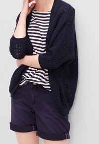 Cárdigan de punto azul marino con tejido abierto, combinado con una camiseta a rayas y pantalones cortos oscuros con dobladillo. Presenta un corte holgado y puños acanalados.
