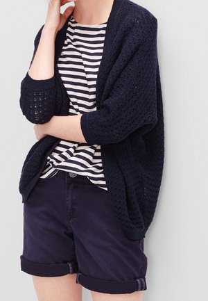 Granatowy sweter z dzianiny o otwartym splotcie, w połączeniu z paskowanym t-shirtem i ciemnymi szortami z podwiniętymi nogawkami. Charakteryzuje się luźnym krojem i ściągaczami na rękawach.