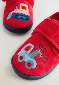 Chaussons en peluche rouge avec des véhicules en cartoon brodés, présentant un tracteur bleu et vert et une pelleteuse jaune, fermés par une sangle en Velcro.