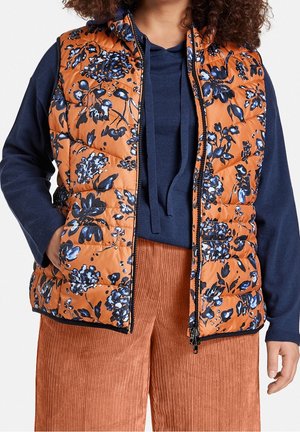 Vrouw draagt een oranje gewatteerd vest met bloemenpatroon over een marineblauwe hoodie met trekkoorden en roestkleurige ribfluwelen broek, handen in de zakken van het vest.