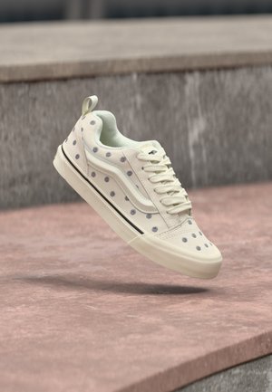 Svetlo krémová nízka teniska s malými modrými kvetinovými vzormi vznášajúcimi sa nad kamennými schodmi.