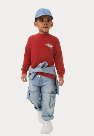 Sweatshirt rouge avec un patch graphique, pull bleu noué à la taille, pantalon cargo baggy bleu clair et baskets blanches. Casquette bleu clair.