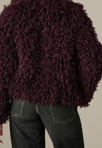 Persona che indossa una giacca texturizzata riccia di un viola scuro e jeans di denim neri, vista da dietro su uno sfondo neutro.