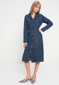 Τζιν μεσαίου μήκους φόρεμα με χελωνίσια λαιμόκοψη, μακριά μανίκια, κουμπιά μπροστά και ταιριαστό ζώνη. Διαθέτει αντίθετη ραφή και σκίσιμο στο στρίφωμα.