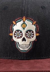 Schwarze Mütze mit einem burgunderroten Wildleder-Schild, das mit einem gestickten Totenkopf-Design mit floralen Akzenten und bunten Details um die Augen verziert ist.