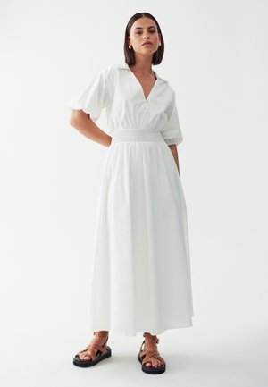 Witte katoenen jurk met korte pofmouwen, v-hals, gerimpelde taille en volle rok. Gepaard met bruine sandalen met bandjes.