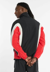 Zwarte jas met rode mouwen, witte accenten, hoge kraag, elastische manchetten en een gestroomlijnd ontwerp. Geschikt voor sport of casual kleding.