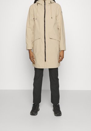 Persoon draagt een beige capuchonjas met voorzakken en rits, zwarte broek en zwarte sneakers, staand tegen een effen lichte achtergrond.