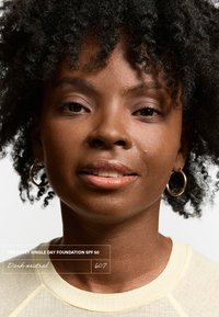 Tube de fond de teint étiqueté "The Every Single Day Foundation SPF 50" en jaune clair avec le texte "Dark neutral 607." Le modèle a des cheveux noirs bouclés et des boucles d'oreilles créoles.