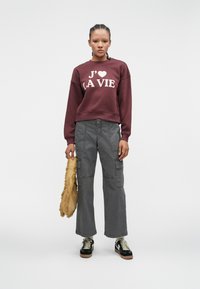 Burgundy sweatshirts with "J'aime la vie" print, gray cargo pants, and black sneakers with white laces. Model holds a fringed tan bag.