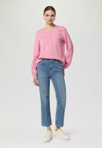 Rosa Langarmbluse mit Schlüssellochdetail, kombiniert mit hellblauen geraden Jeans und beigen Sneakers, vor einem neutralen Hintergrund.