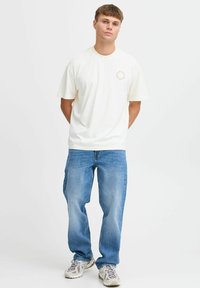Cream-colored cotton t-shirt with short sleeves and a round neckline, featuring a small graphic design on the chest, paired with blue jeans.