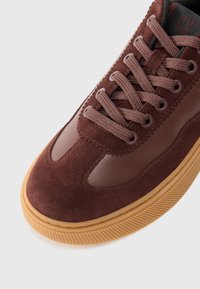 Brown suede sneaker with a smooth leather panel, round toe, textured laces, and a gum rubber sole featuring horizontal ridges.