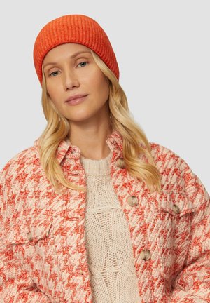 Un bonnet rouge côtelé porté avec un pull texturé crème et une veste à motif pied-de-coq rose et blanc, ornée de boutons ronds et offrant un look confortable.
