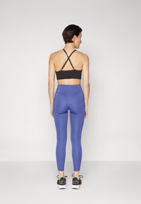 Svart sports-bh med kryssrygg, kombinert med høy midje blå leggings. Modell iført svart og hvit treningsko. Nærbilde.