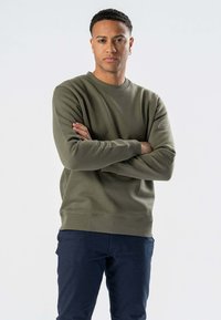 Oliven grøn sweatshirt lavet af en bomuldsblanding, med rund halsudskæring, ribmanchetter og ribkant. Båret med mørkeblå bukser, med armene korslagte.