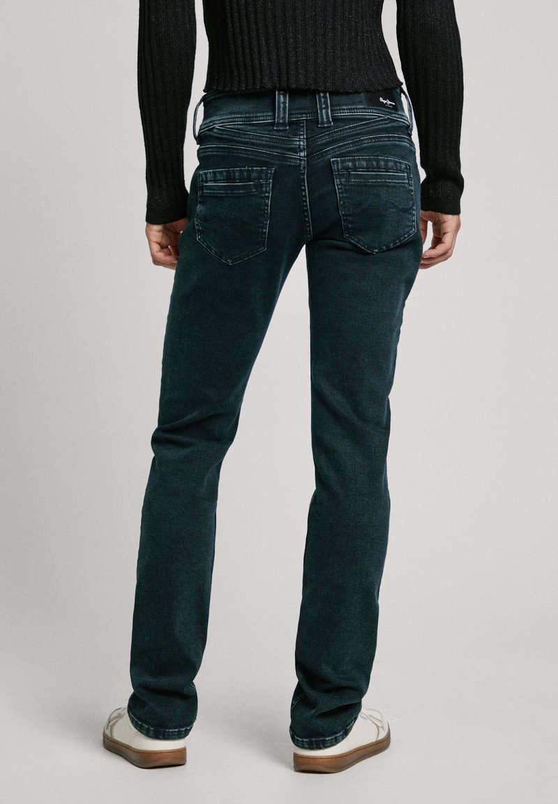 Dunkelgrüne Jeans mit geradem Bein, leicht taillierten Gesäßtaschen und dezentem Nähdetail. Kombination mit einem schwarzen gerippten Pullover und weißen Schuhen.