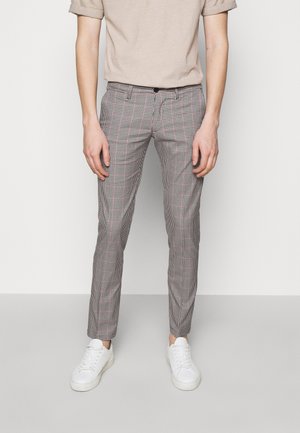 Person iført tætsiddende ternede bukser, beige kortærmet skjorte og hvide sneakers, stående mod en ensfarvet baggrund.