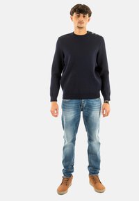 Maglione lavorato a maglia blu navy con orlo e polsini a costine, caratterizzato da dettagli con bottoni sulla spalla. Abbinato a jeans chiari e stivali marroni con lacci.