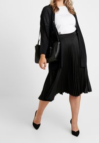 Černá skládací sukně s texturovaným černým kardigánem a bílým tričkem. Kombinováno s černými lodičkami s úzkou špičkou a strukturovanou černou kabelkou.
