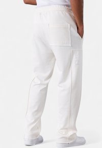 Person, der beige Sweatpants mit zwei sichtbaren Taschen trägt, wobei eine mit einer Biene bestickt ist, kombiniert mit weißen Sneakers und einem weißen Oberteil.