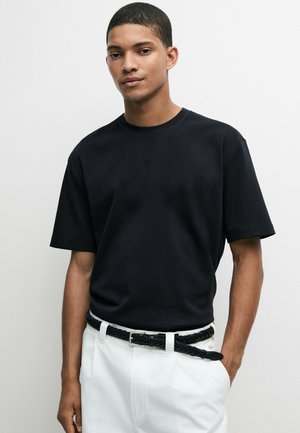 Ung mand med kort hår iført en sort kortærmet T-shirt og hvide bukser med sort flettet bælte mod en ensfarvet baggrund.