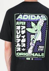 T-shirt noir avec un design graphique aux accents verts et violets. Le texte comprend "ADIDAS", "SUPER" et une illustration de personnage de dessin animé ludique.