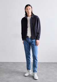 Námořnický zipový svetr, krémová košile, světle modré vzorované džíny a bílé tenisky. Model se fotí na neutrálním pozadí.
