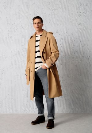 Beige Trenchcoat mit Doppelreiher-Design, kombiniert mit einem schwarz-weißen gestreiften Hemd, grauen Jeans und dunkelbraunen Stiefeln. Texturierte Wand im Hintergrund.