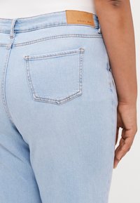 Light wash denim jeans with a high waist, a textured finish, and a brown leather patch labeled "NOISY MAY" at the back waist.