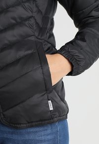 Doudoune noire avec un design matelassé, des poignets élastiques et une poche latérale. Fabriquée à partir d'un matériau lisse et brillant avec une étiquette de logo discrète.