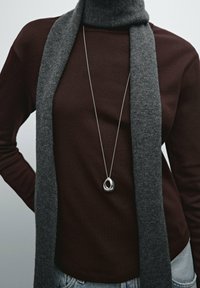 Gray scarf worn over a brown long-sleeve top, featuring a silver necklace with a rounded, abstract pendant. Denim jeans visible at the bottom.