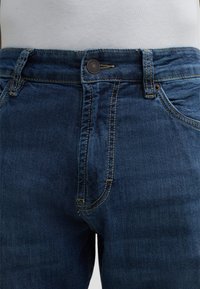 Granatowe dżinsy o klasycznym pięciopunktowym fasonie, z widocznym szyciem, metalowym zapięciem i gładką fakturą.