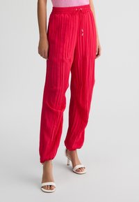 Pantaloni di stoffa rossi, stropicciati, con vita elastica, stringa regolabile e polsini alla caviglia. Abbinati a sandali bianchi con tacco.