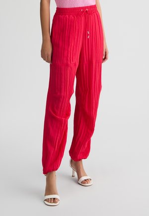 Pantaloni di stoffa rossi, stropicciati, con vita elastica, stringa regolabile e polsini alla caviglia. Abbinati a sandali bianchi con tacco.