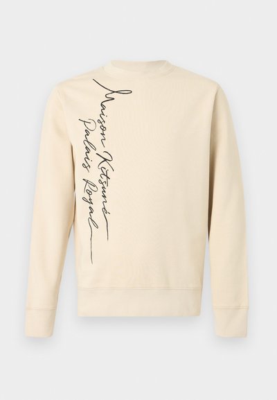 Μακρυμάνικη μπλούζα σε κρεμ χρώμα με κατακόρυφο μαύρο γραφικό κείμενο σε καλλιγραφικό στυλ που γράφει "Maison Kitsuné Palais Royal" στην αριστερή πλευρά.
