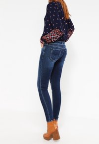 Pantalones ajustados de mezclilla de color azul, con una cintura de altura media y un diseño de cinco bolsillos. Llevados con una blusa de manga larga estampada y botas marrones de tacón.