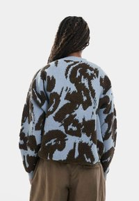 Personne aux longs cheveux tressés portant un pull à motifs bleu et marron et un pantalon marron, vue de dos sur un fond uni.