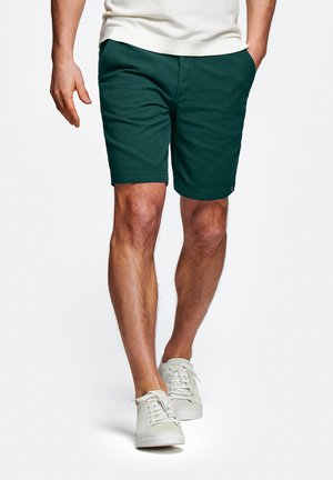 Man loopt en draagt donkergroene korte broek, witte sneakers en een wit shirt met korte mouwen, met één hand in zijn zak.