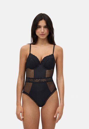 Bodysuit de lencería negra con copas moldeadas, acentos de encaje, paneles de tela transparente y tirantes delgados ajustables. Piernas de corte alto y bordes scalloped.