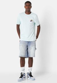 T-shirt di cotone azzurro chiaro con grafica sul petto, abbinato a pantaloni corti in denim strappati e sneakers alta nere e blu.